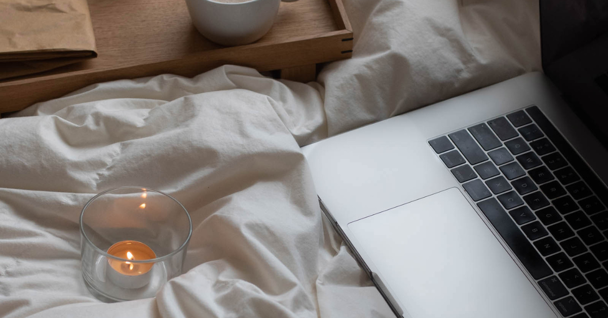 Laptop, candle, and tray with coffee cup on top of white bed sheets